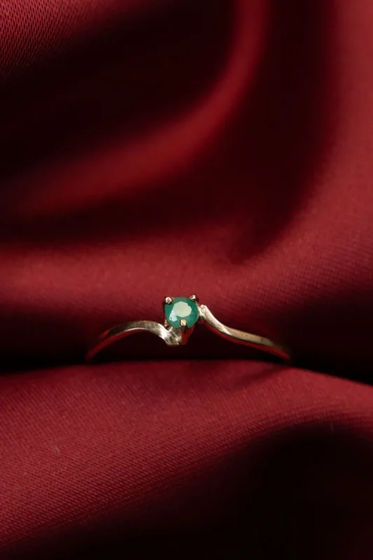 Anillo delicado de oro 18k con una pequeña esmeralda verde sobre tela roja elegante.