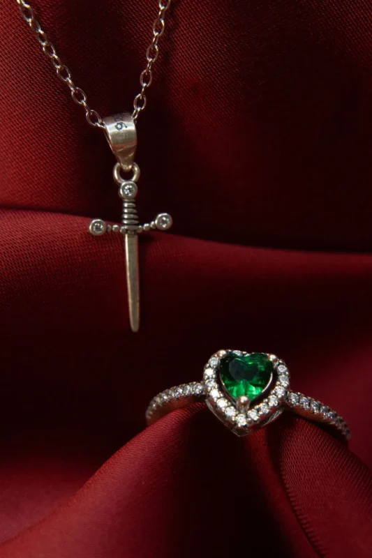 Anillo de plata ley 925 con circones, diseño único en forma de corazón verde rodeado de circonitas, acompañado por un adorno en forma de espada, sobre fondo de tela roja.