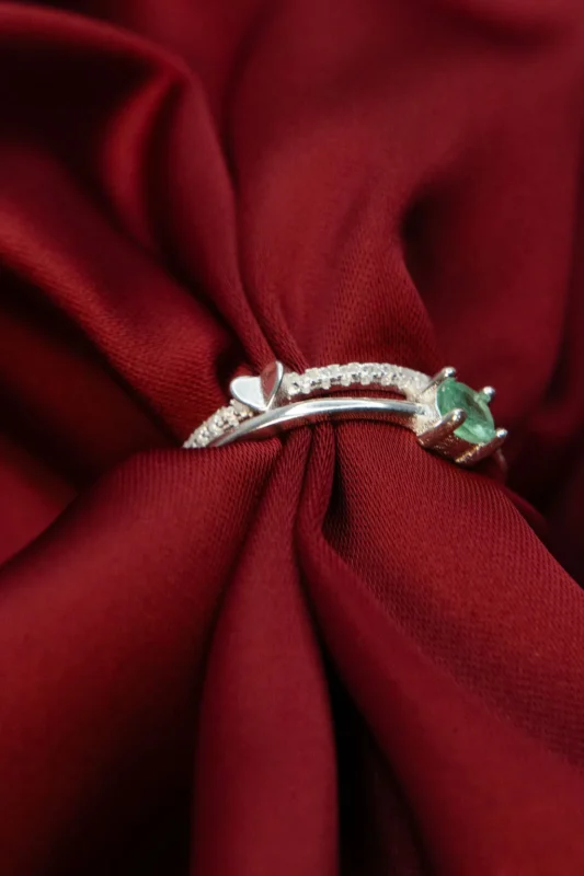 Anillo de plata ley 925 con esmeralda y circones, diseño único con detalle de corazón y engaste lateral, fotografiado sobre tela roja.