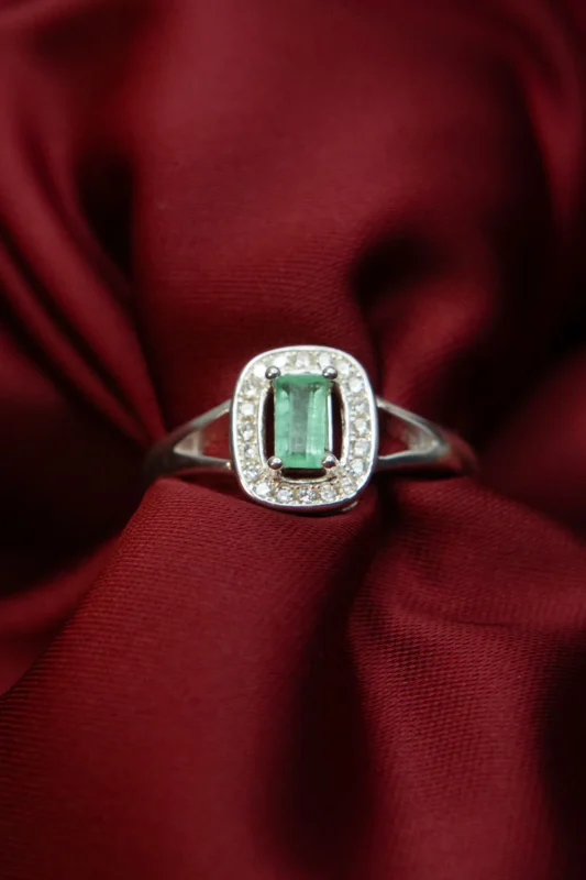Anillo de plata ley 925 con una esmeralda central de corte rectangular y rodeado de circones, diseño único, posado sobre tela roja.
