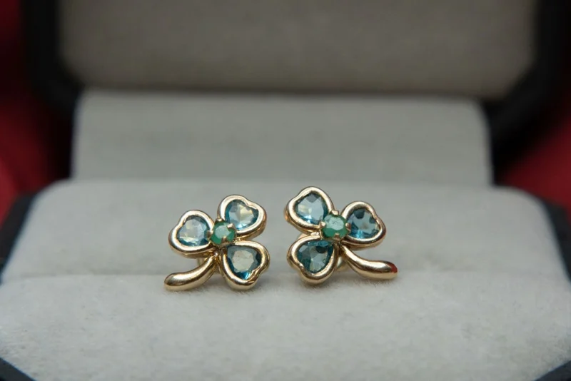 Aretes en oro de 18K en forma de trébol, con esmeraldas y circones, sobre fondo de estuche gris.