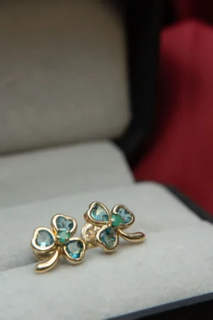 Aretes en oro de 18K en forma de trébol, con esmeraldas y circones, sobre fondo de estuche gris.