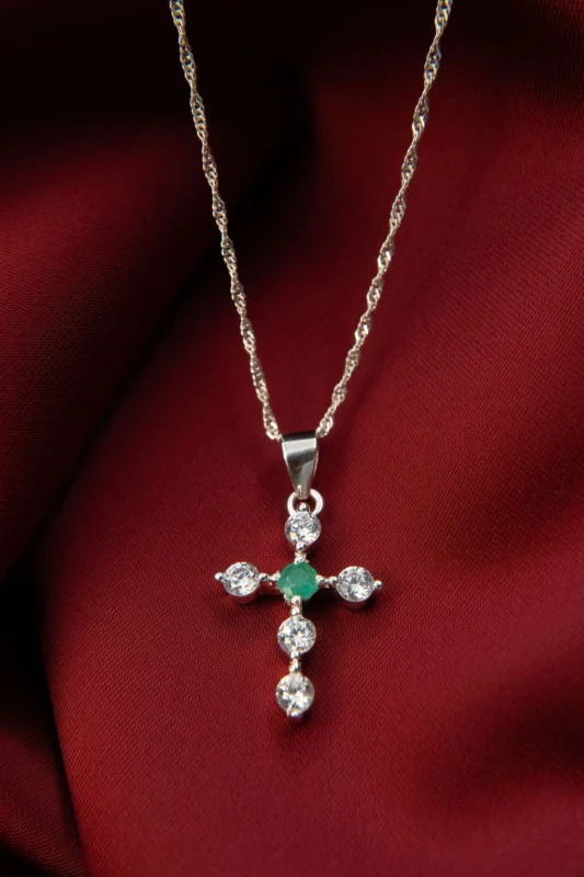 Collar de plata ley 925 con dije en forma de cruz adornado con circones y una esmeralda central, sobre fondo de tela roja.