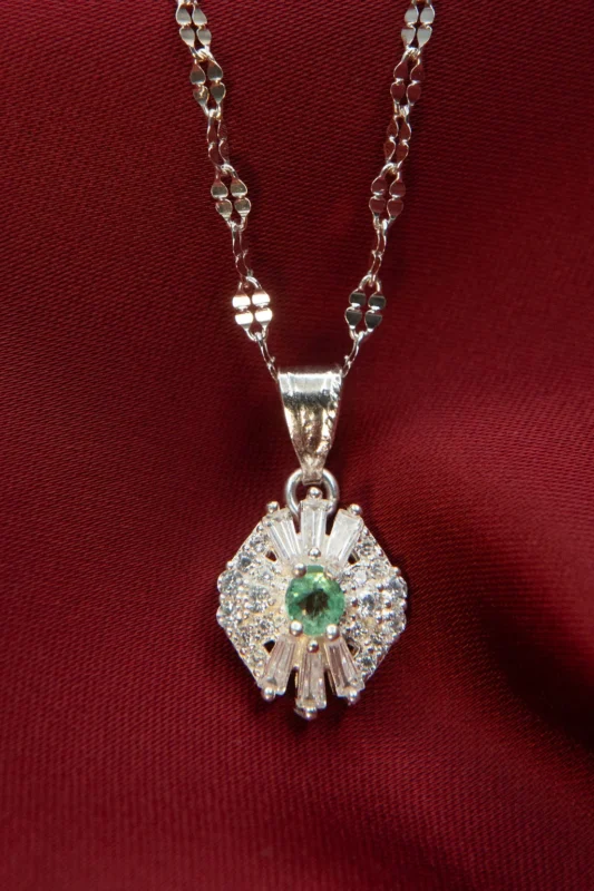 Dije en plata ley 925 con circones y esmeralda natural, diseño hexagonal con piedra central verde, fotografiado sobre tela roja.