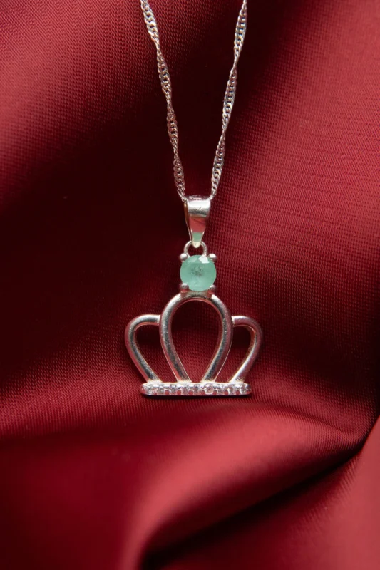Collar con dije de corona en plata ley 925, decorado con circones y esmeralda natural sobre fondo rojo.