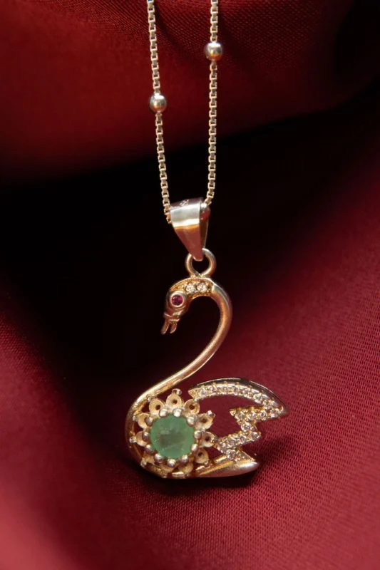 Dije en forma de cisne elaborado en plata ley 925, decorado con circones y esmeralda natural, sobre fondo de tela roja