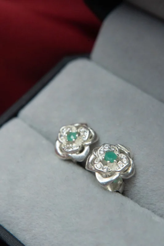 Aretes en plata ley 925 con diseño de flor, esmeralda en el centro y circones, presentados en estuche gris.