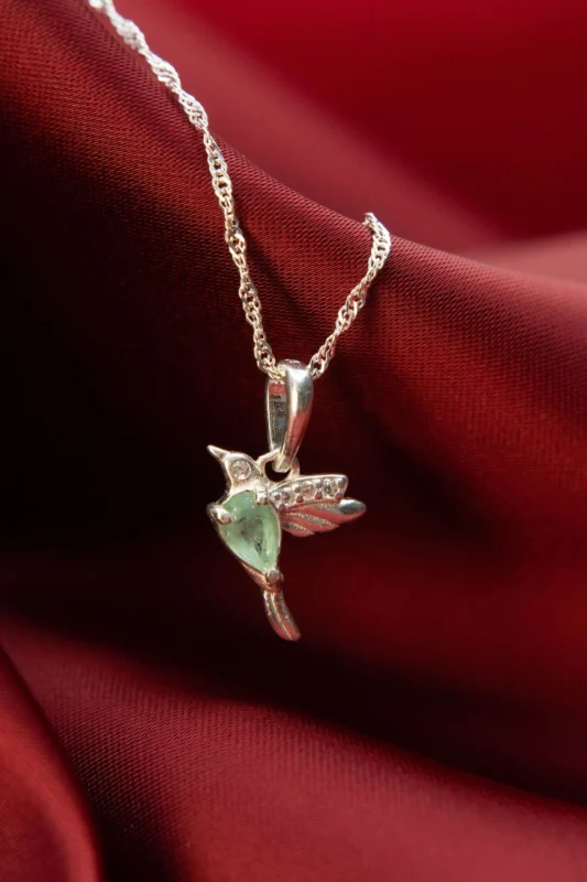 Dije en plata ley 925 con forma de colibrí, decorado con circones y esmeralda natural, sobre fondo de tela roja.