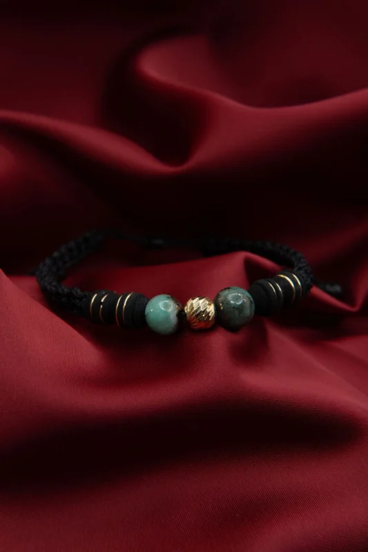 Pulsera en hilo negro con balines de esmeralda natural y balines de oro 18K, presentada sobre fondo de tela roja.