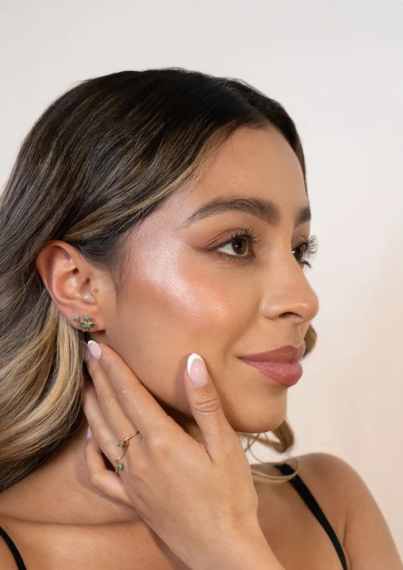 Primer plano de mujer con aretes y anillo de oro 18K con esmeraldas y circones, sobre fondo neutro.