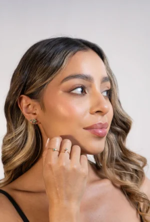 Primer plano lateral de mujer con cabello suelto, luciendo aretes de oro 18K con esmeraldas y circones, en fondo neutro.