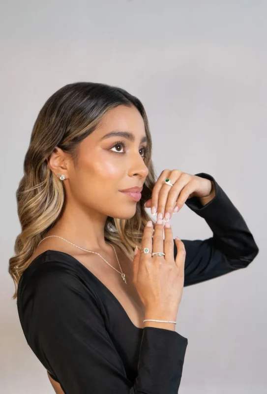 Mujer de perfil con cabello suelto y vestido negro, luciendo aretes, collar colibrí, pulsera y anillos en plata ley 925 con esmeraldas y circones.