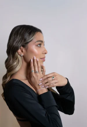 Mujer con cabello suelto y vestido negro mostrando aretes y anillos de plata ley 925 con esmeraldas y circones, sobre fondo neutro.